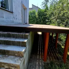 Nowy, wysoki taras na słupach, z betonowymi schodami prowadzącymi do niego. Otoczony zielenią.