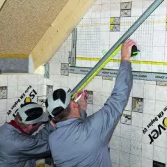 Dwóch mężczyzn pracuje przy montażu grzybków na poddaszu, mierząc i zaznaczając punkty na folii paroizolacyjnej.