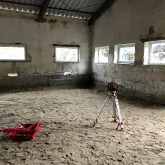 Na piaszczystym podłożu stoi niwelator geodezyjny, gotowy do pracy. W tle widać okna i ściany, a na ziemi leży czerwona skrzynka. To przygotowania do wylania posadzki z **gruntu do anhydrytu**.