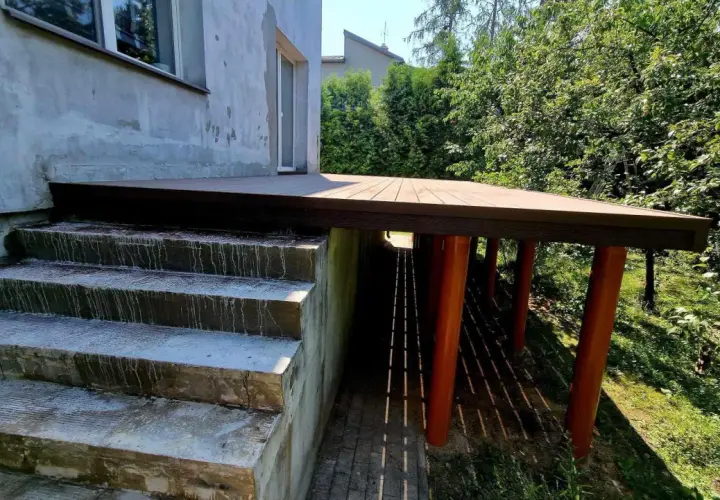 Nowy, wysoki taras na słupach, z betonowymi schodami prowadzącymi do niego. Otoczony zielenią.
