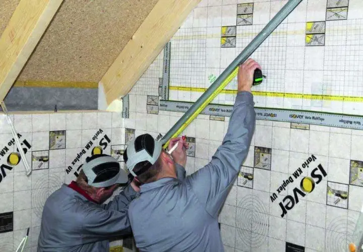 Dwóch mężczyzn pracuje przy montażu grzybków na poddaszu, mierząc i zaznaczając punkty na folii paroizolacyjnej.