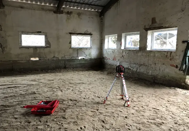Na piaszczystym podłożu stoi niwelator geodezyjny, gotowy do pracy. W tle widać okna i ściany, a na ziemi leży czerwona skrzynka. To przygotowania do wylania posadzki z **gruntu do anhydrytu**.