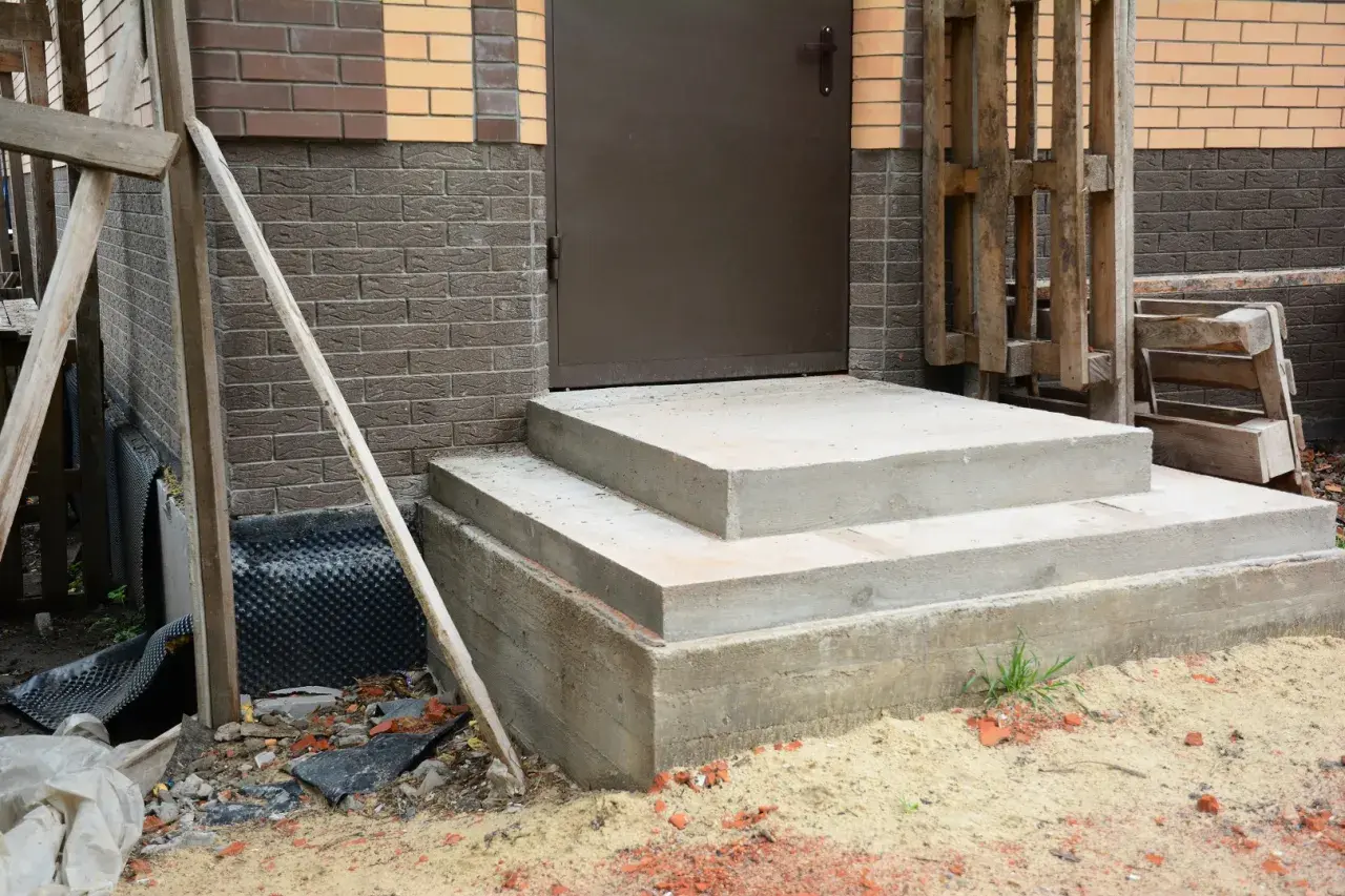Solidny fundament pod schody z betonu, gotowy na dalsze prace budowlane. Obok widać materiały budowlane i fragment domu.