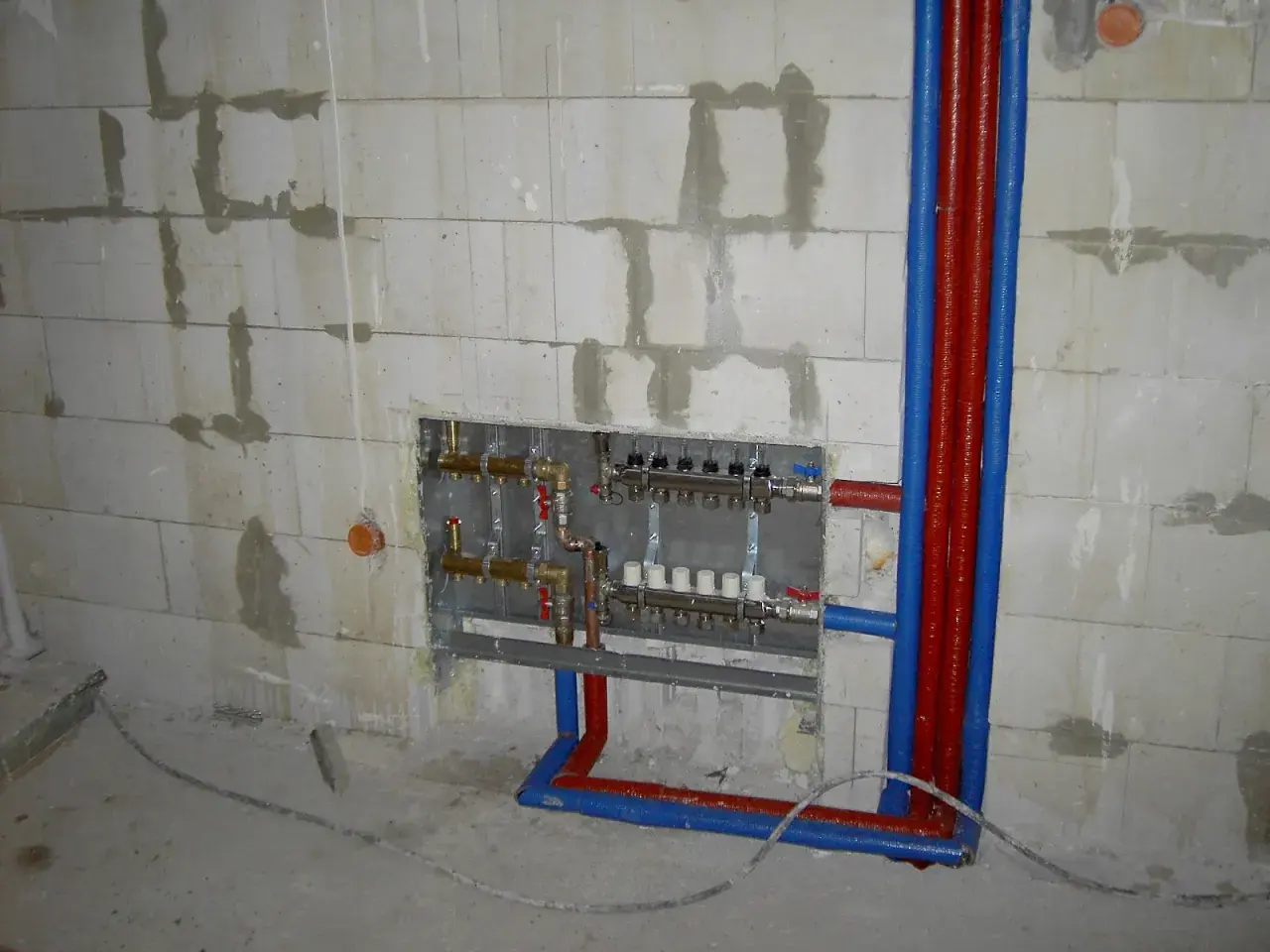 Instalacja hydrauliczna przed tynkami: widoczny rozdzielacz ogrzewania podłogowego i rury w ścianie z bloczków.