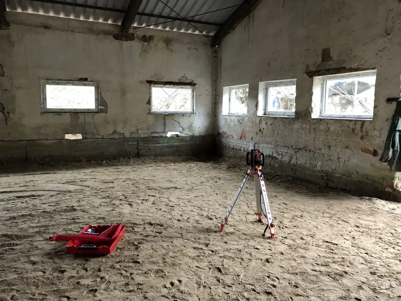 Na piaszczystym podłożu stoi niwelator geodezyjny, gotowy do pracy. W tle widać okna i ściany, a na ziemi leży czerwona skrzynka. To przygotowania do wylania posadzki z **gruntu do anhydrytu**.