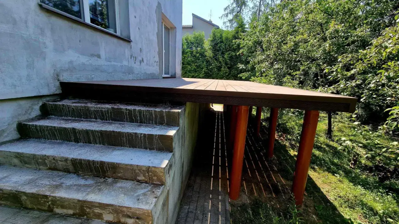Nowy, wysoki taras na słupach, z drewnianymi stopniami prowadzącymi do domu. Otoczony zielenią.
